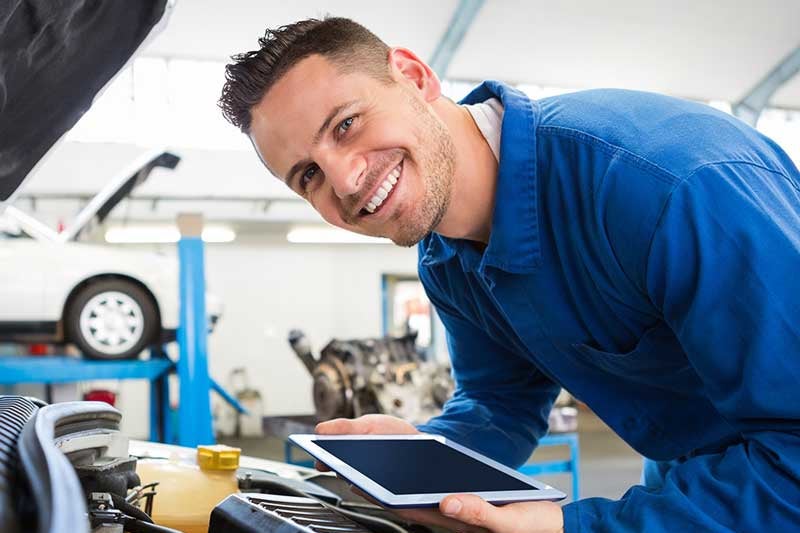 A blue-shirted certified mechanic inspecting a vehicle engine in an automotive service center looks up and smiles encouragingly.